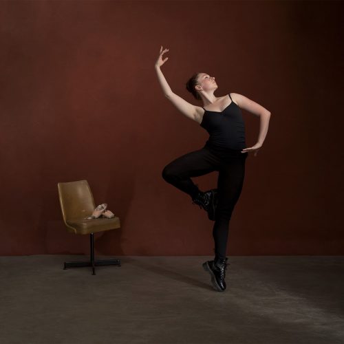 Ballet dancer in black tights and leotard posing in Doc Martins while ballet shoes sit on the chair in the background.