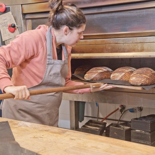 A baker eases four loaves of bread out of the oven with a timber peel