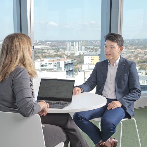 Two people having a meeting in a corporate environment, sitting near large glass windows showing the urban sprawl, and one person has a laptop in front of them