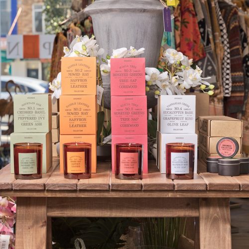 Four varieties of scented candles are stacked in rows on a timber table. Each variety has a candle on display for smelling.